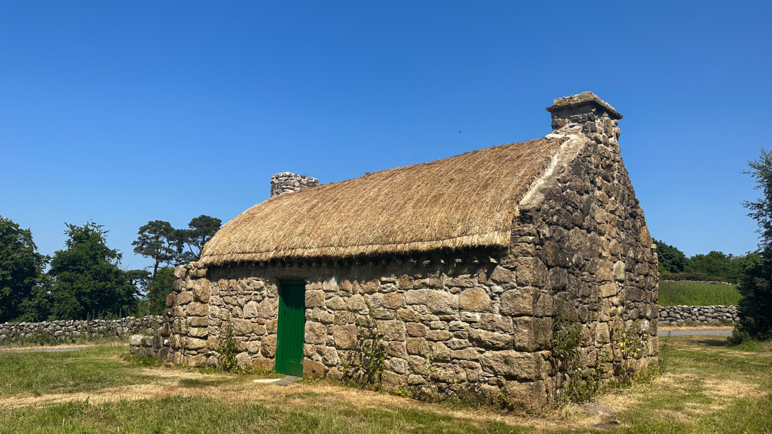A small thatched cottage sits in a clear. The sky is blue and sunny.