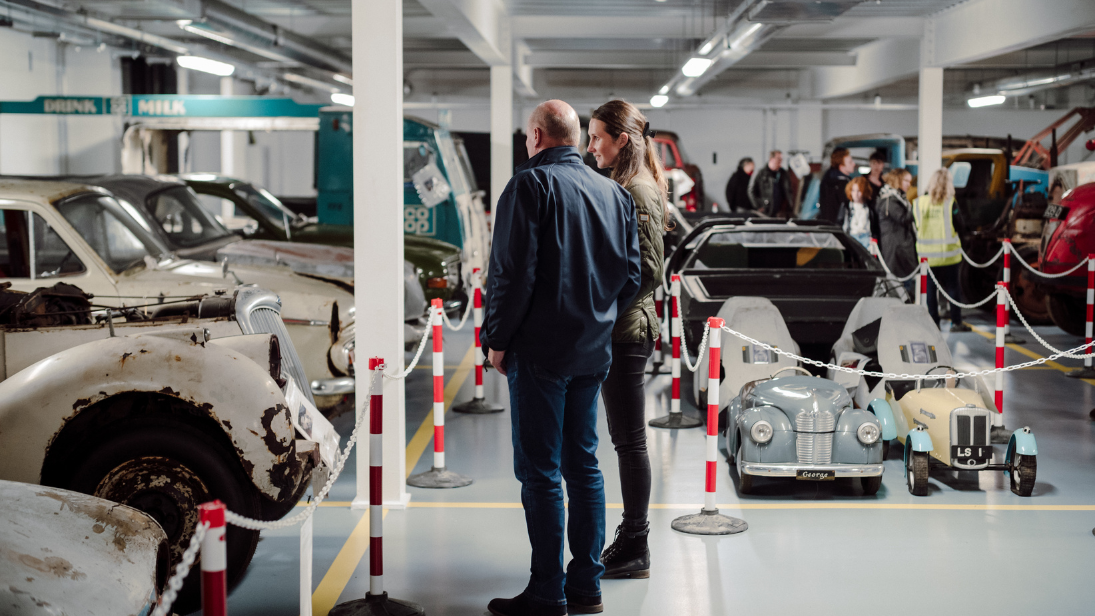 Two people standing looking at a car in the closed off collection of the Transport and Industry Store of the Ulster Transport Museum. More collection cars and people in the background.