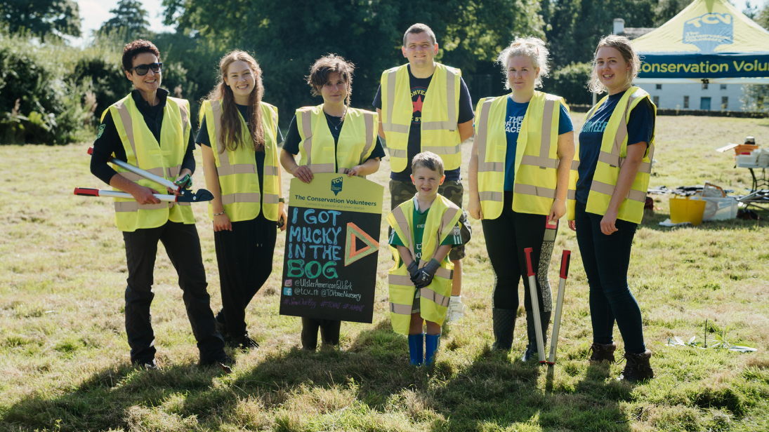 A group of volunteers in high vis vests, mix of ages, at the bog site at the Ulster American Folk Park in Omagh. One volunteer is holding a sign saying I got mucky in the bog with The Conservation Volunteers logo on it and a gazebo with the same logo on it is in the background.