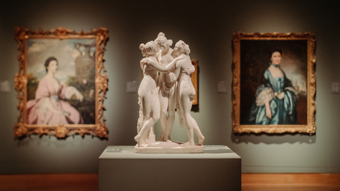 a small white sculpture of three people embraced and two paintings in the background