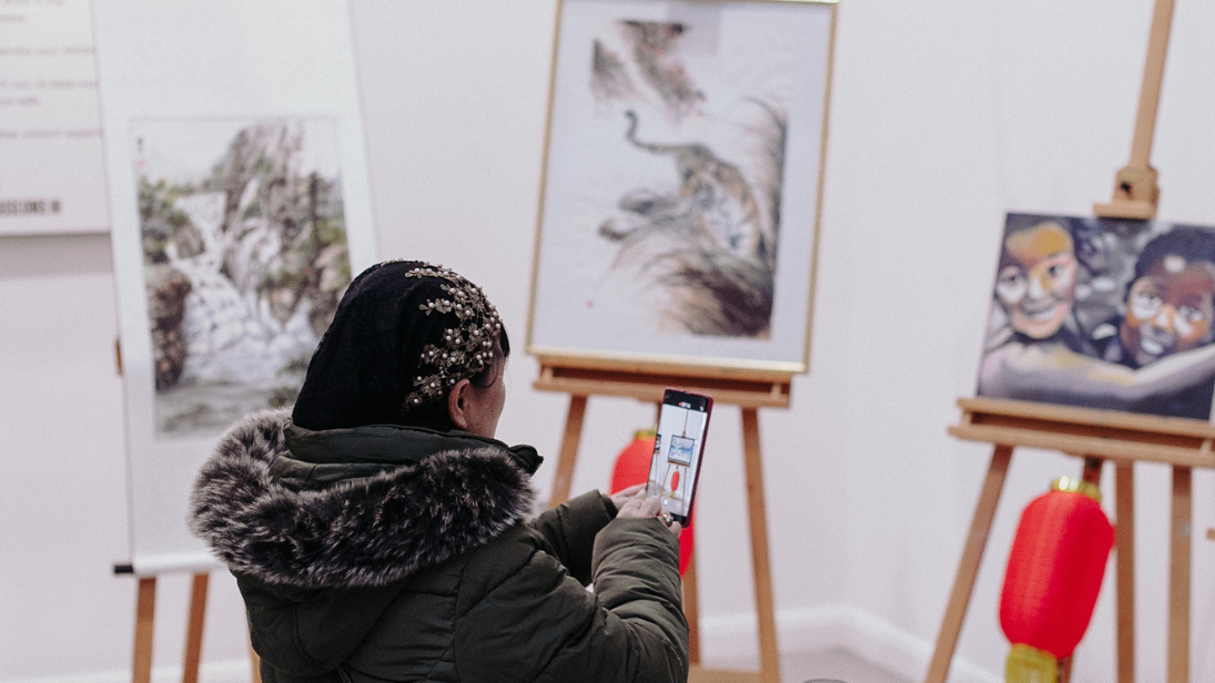 A woman in a black coat looking at Chinese paintings and taking a photo of the paintings on her phone. Red Chinese lanterns hang from the eassels.