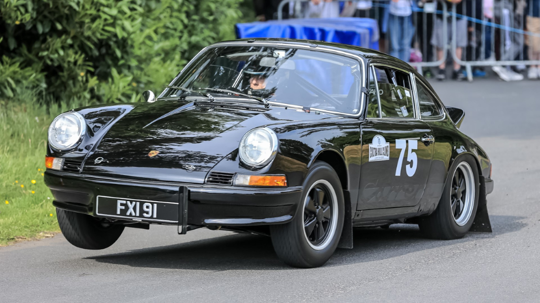 A black Porsche being driven by a rally driver wearing a helmet up a hill with crowds behind a barrier pictured in the background. 
