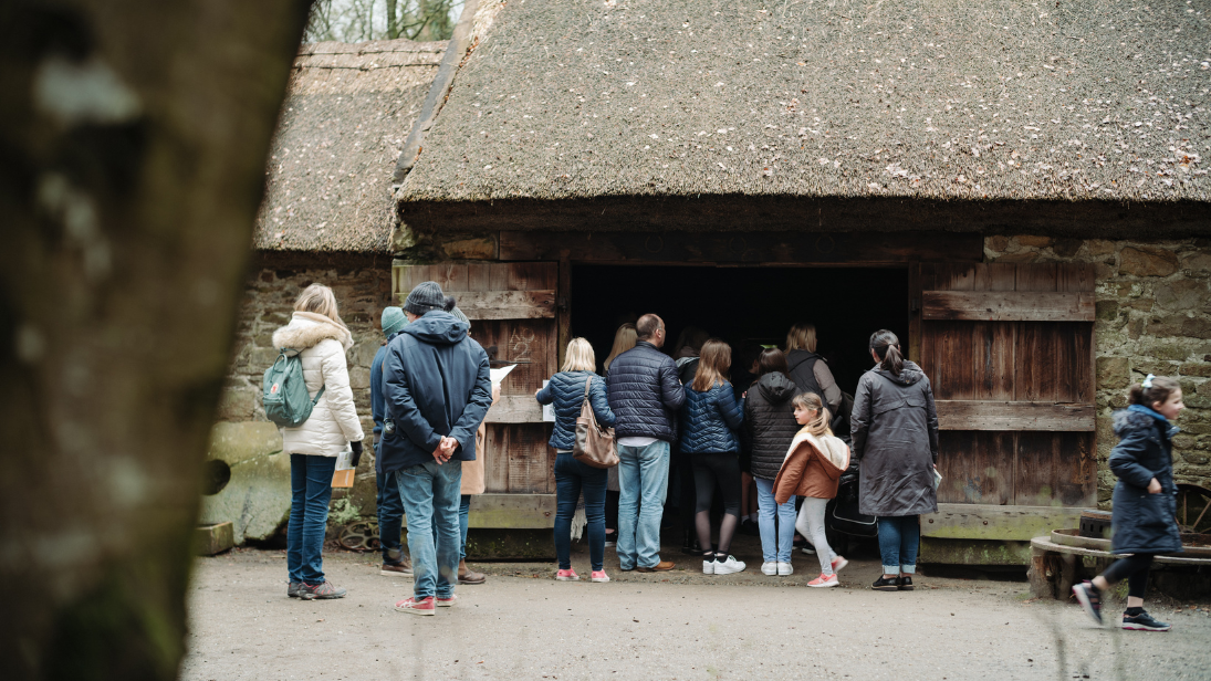 A group of people, mixture of adults and kids, huddled around the opening to an authentic, Ulster stone-built dwelling in the Ulster American Folk Park. 