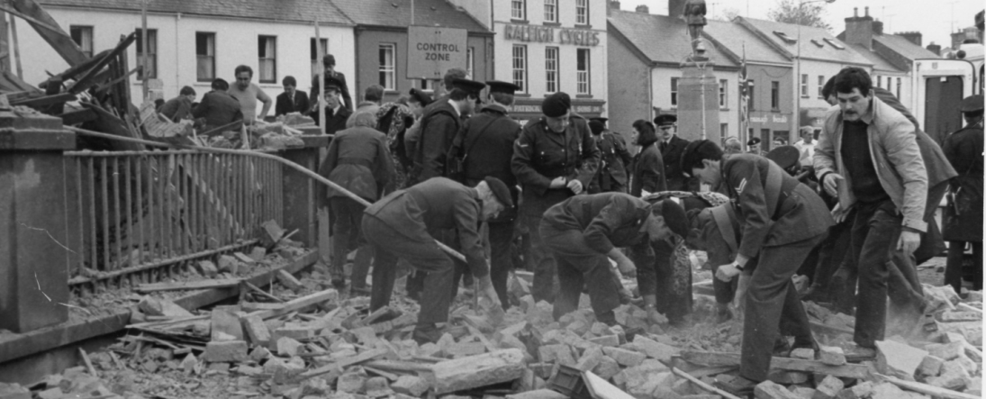 Black and white image of the aftermath of the Enniskillen bomb. Men and police officers are moving rubble.