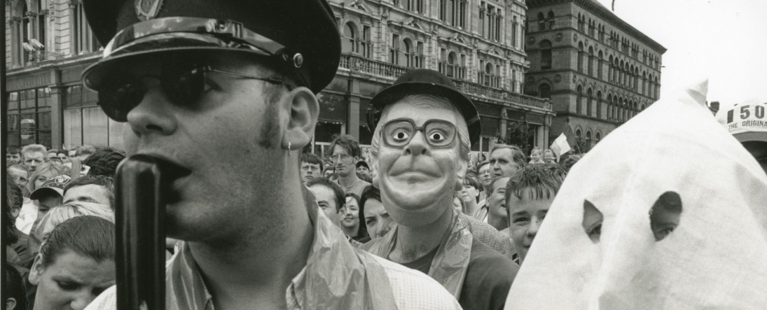 Black and white photograph of the Drumcree protest at Belfast City Hall. Man in the forefront wearing sunglasses, hat and holding a baton to his mouth. Featured person wearing a mask and hat in the background, and person under a white sheet with the eyes cut out in the foreground