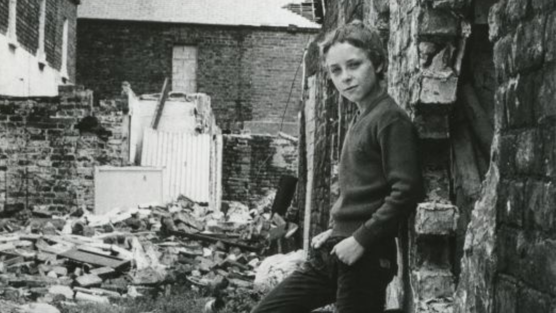 Black and white photograph of a young boy leaning against a wall, background is the aftermath of a bomb.