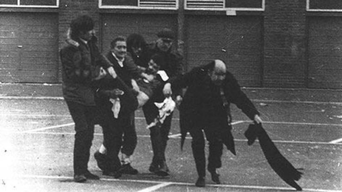 Black and white photograph of an injured person being carried, one person is waving a white hanky