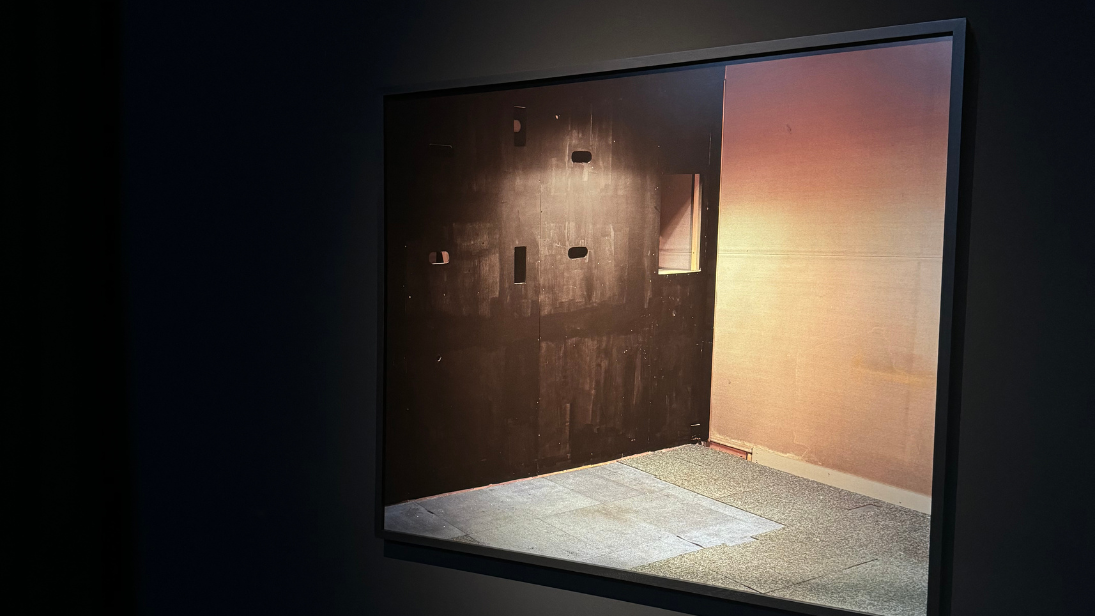 A picture of a wooden art installation, which is a wall with holes cut into it, in a black frame on a black wall.