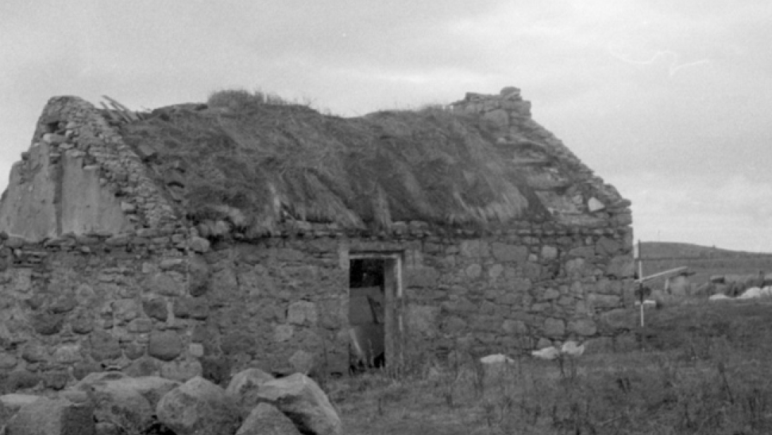A black and white image of a derelict cottage with a thatched roof falling in and no door.