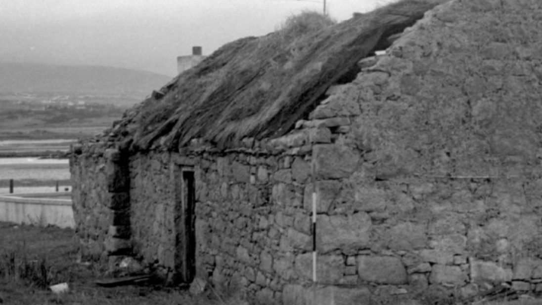 A black and white image of a derelict cottage with a thatched roof falling in. In the background we can see a body of water. 