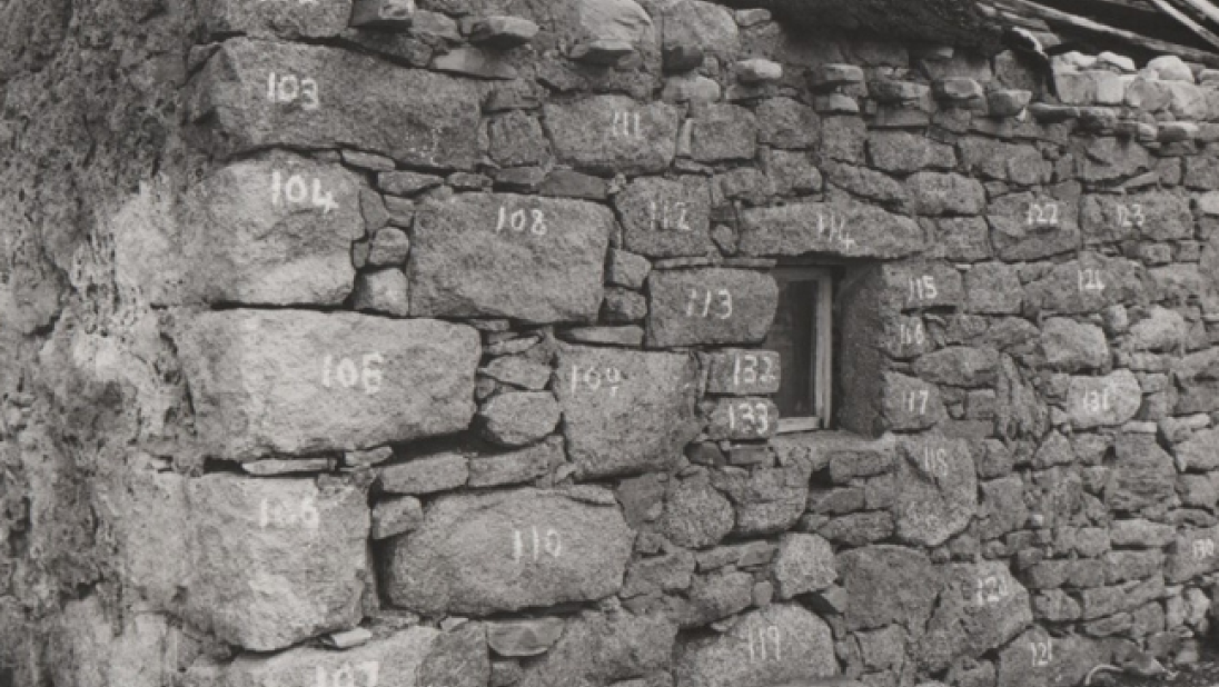 A black and white image of the walls of a run-down cottage. The stones are numbered with white chalk. 
