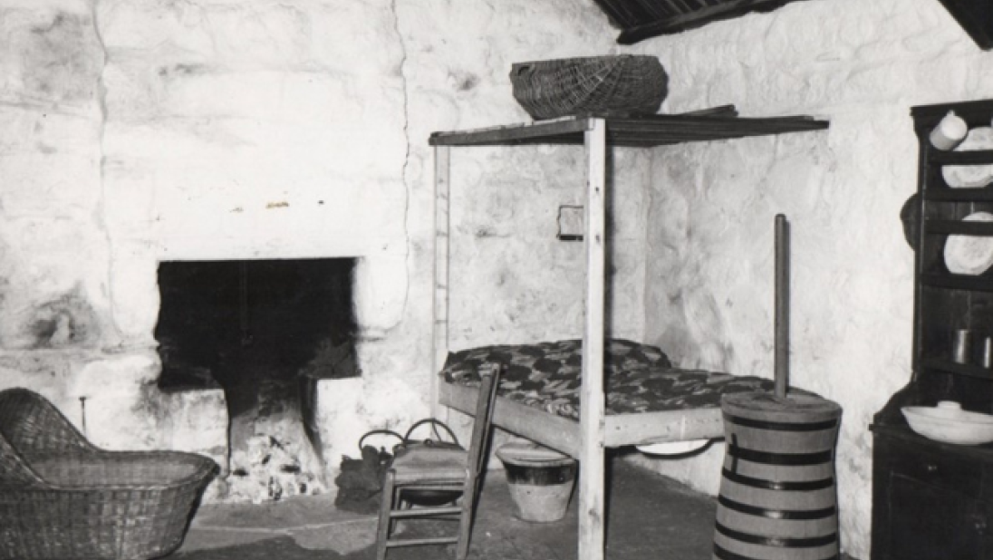The inside of a byre dwelling staged with objects including a butter churn, a corner bed, a dresser with crockery, and a baby's cradle. 