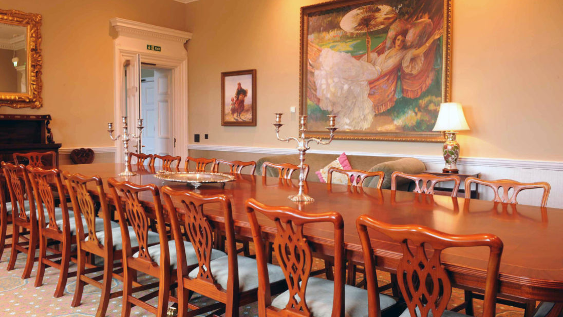 Long table and chairs in a dining room with two candelabras up the middle of the table. A large, colourful painting is on the wall behind and a lit lamp on the side table.