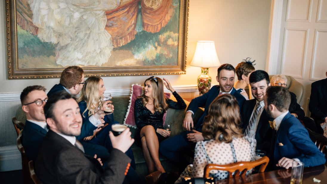 A group of smartly dressed people sitting between a sofa and chairs at a party talking and enjoying themselves. There is a painting in the background of the picture and a lit lamp.