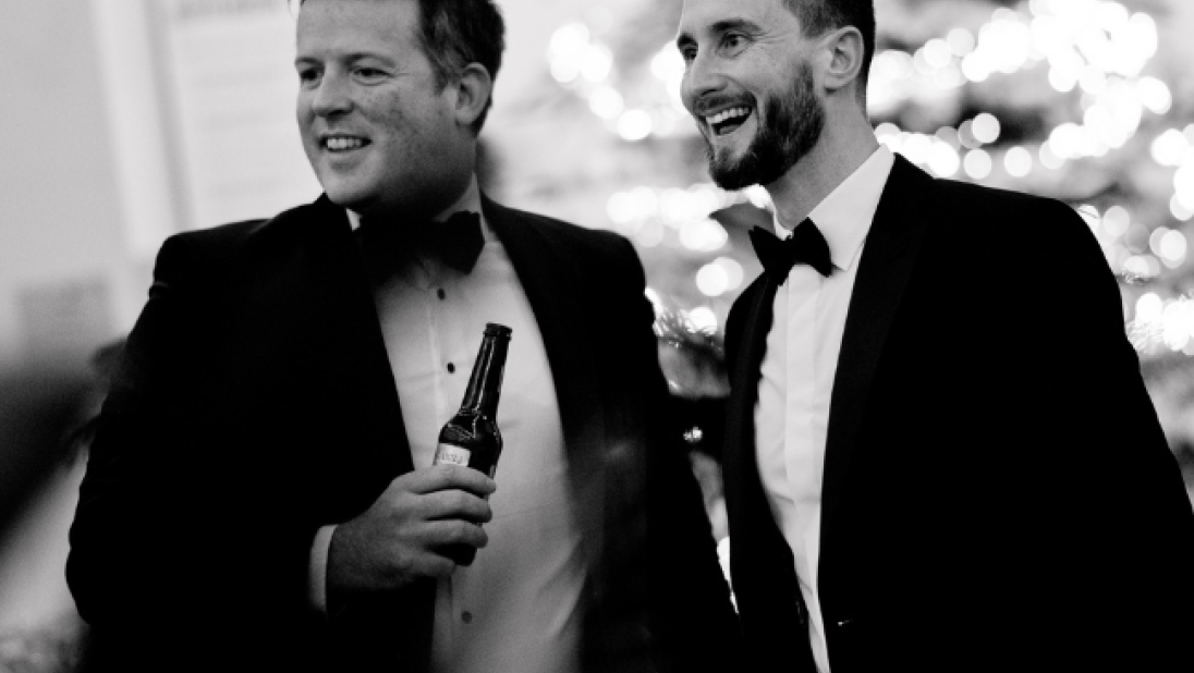 A black and white image of two men dressed in tuxedos, one is holding a bottle of beer, with a lit up Christmas tree in the background.