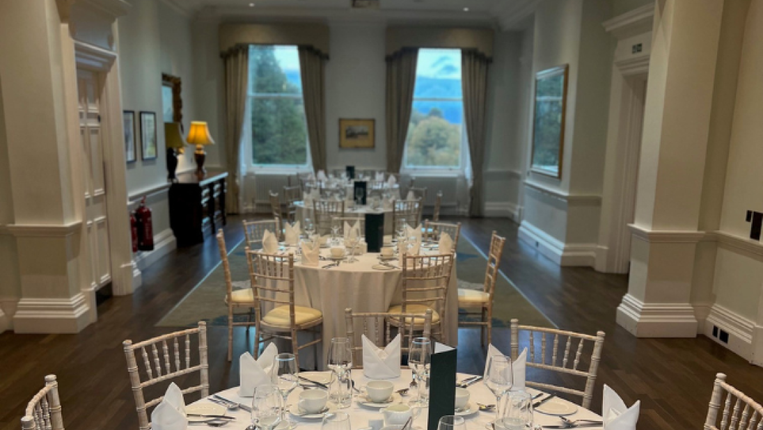 Three round tables laid out in the drawing room of Cultra Manor, all dressed in white cloth. Two large windows in the background let in natural light and a chandelier hangs above one of the table.