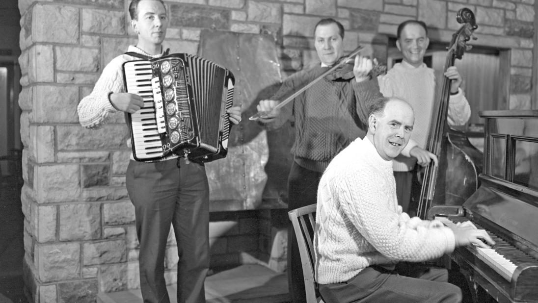 Four musicians pose for a photo. They are playing an accordian, a piano, a fiddle, and what appears to be an upright bass.