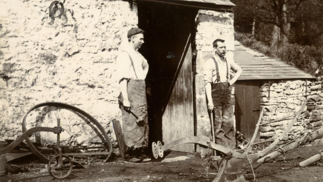 Two blacksmiths stand outside a forge, with many metal objects for agriculture around them.