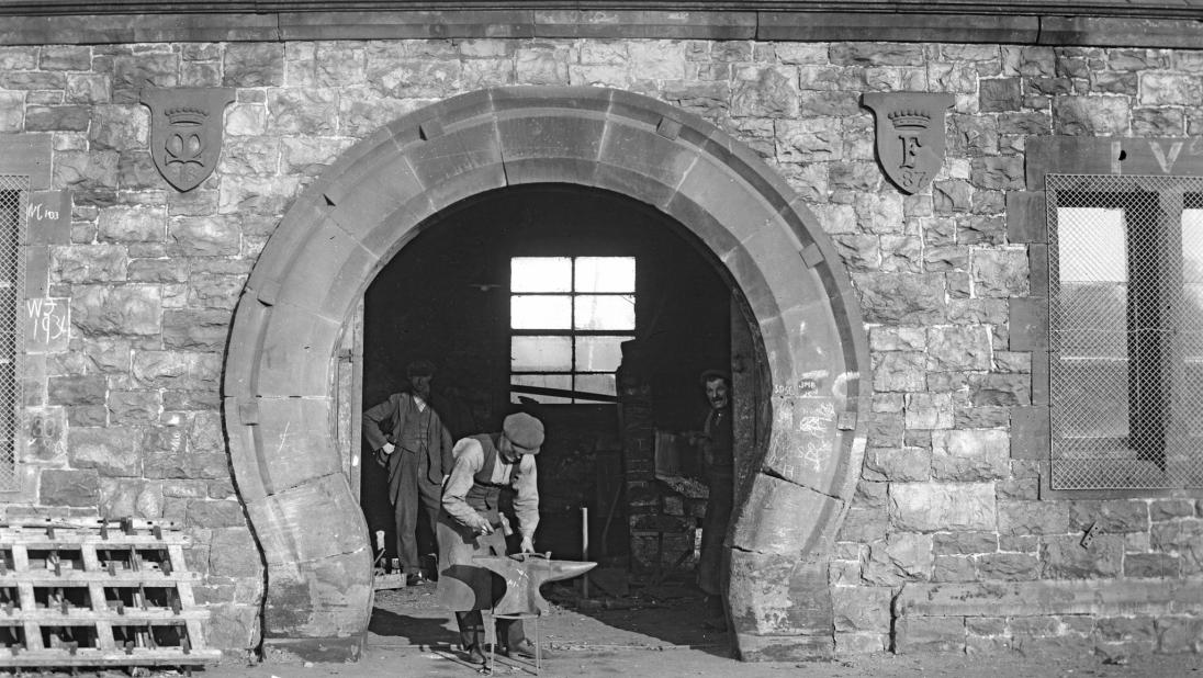 The entrance to a forge, with a man inside staring out. Another man in the doorway is working on an anvil.