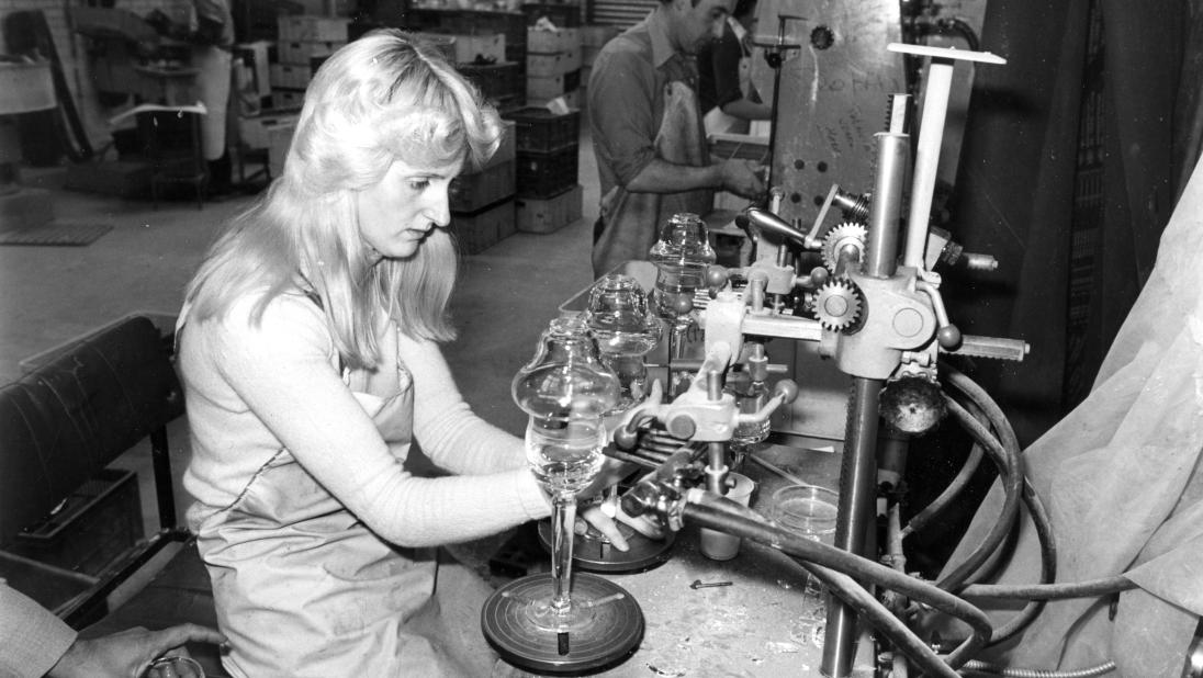 A woman sits at a work desk, working with blown glass.