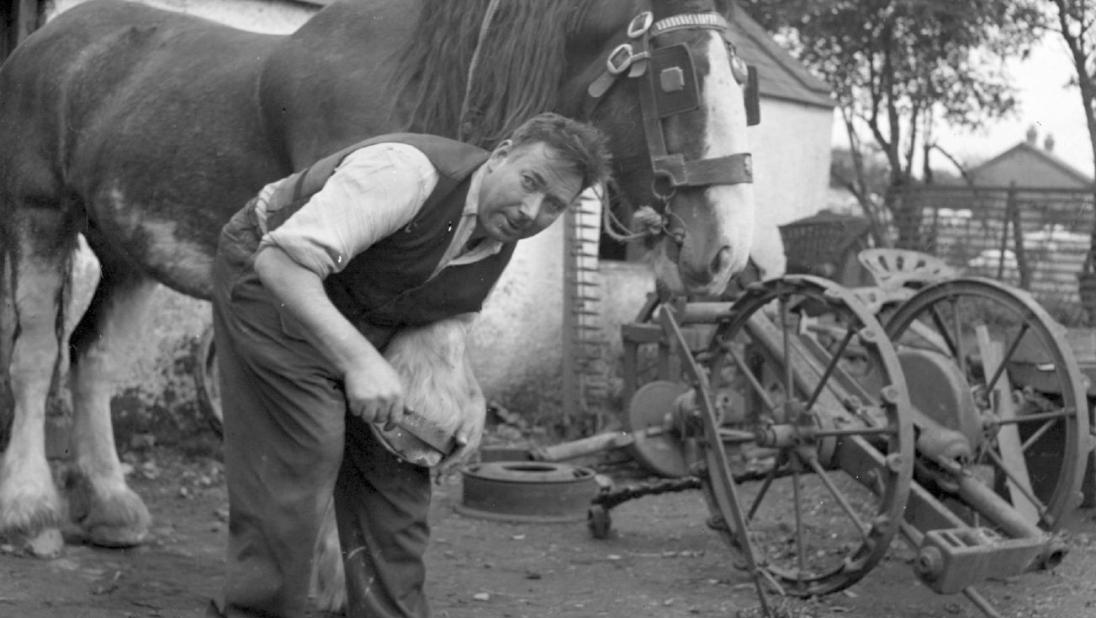 A black and white image of a man looking towards the camera as he shoes a horse.