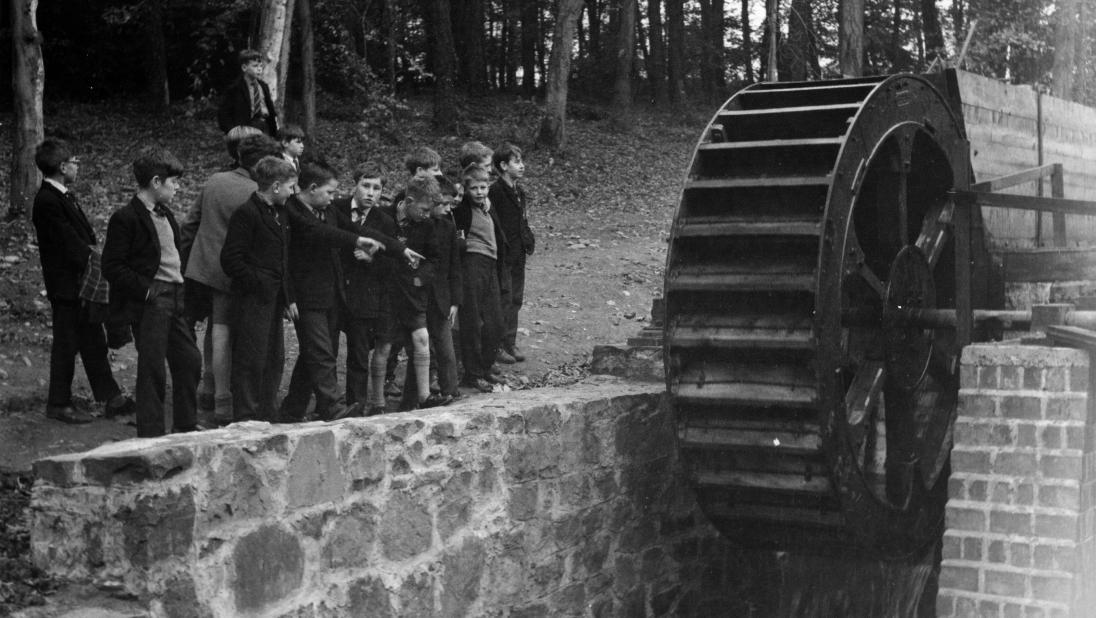 A black and white image of a school group looking at a water wheel.