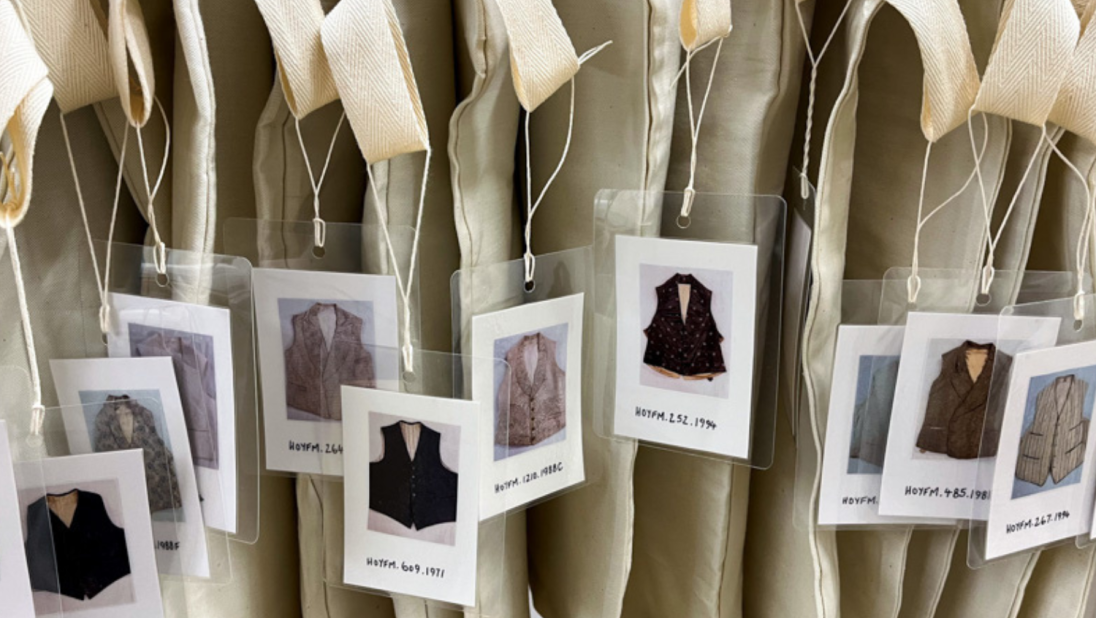 A row of waistcoats wrapped in white covers with labels dangling outside.