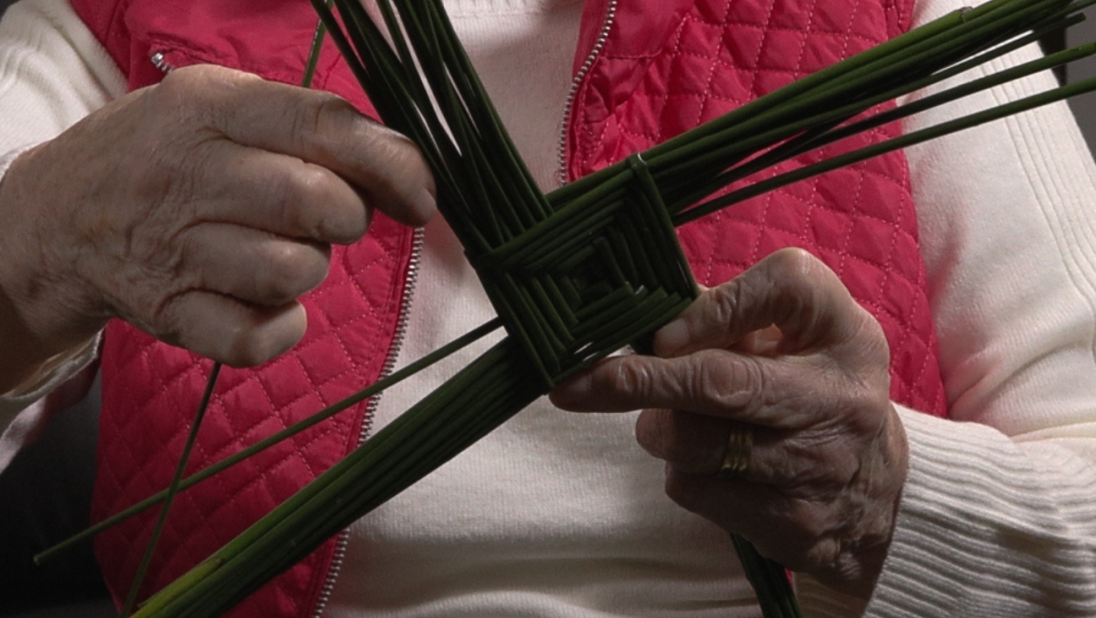 A woman in a red cardigan weaves a St. Brigid's Cross.