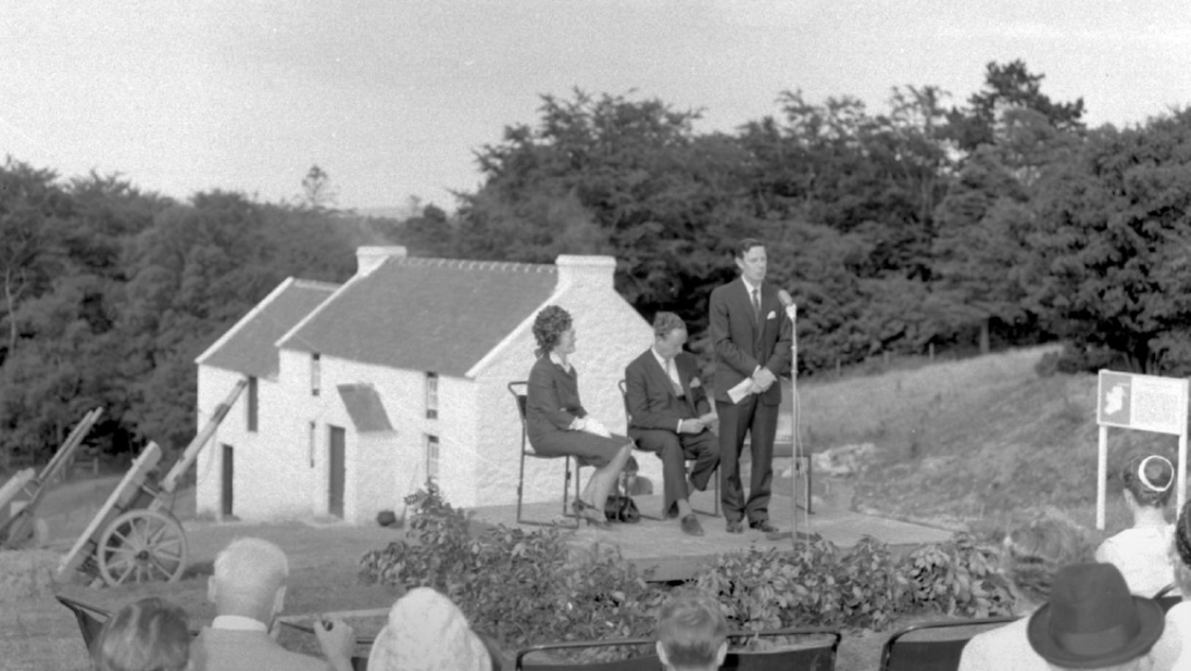 A black and white image of an event. In the foreground, a group of people are listening to a speaker on a podium. In the background, a farm sits on a sloping hill.