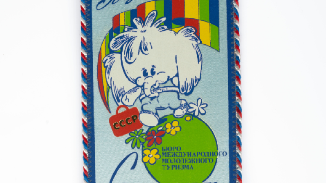 A small banner. The edges are in rainbow thread. The banner is light blue with images and cyrillic text. The image is a cartoon elephant on a green globe with flowers, holding a red briefcase with USSR written on it, and a rainbow background.