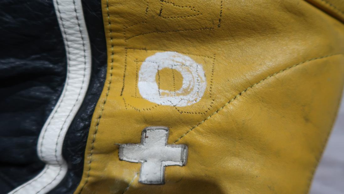 A close up of the top of a racing jumpsuit. There are white patches spelling 'O +' sewn on. The patches look like they are more closely adhereing to the jumpsuit than in previous photographs.