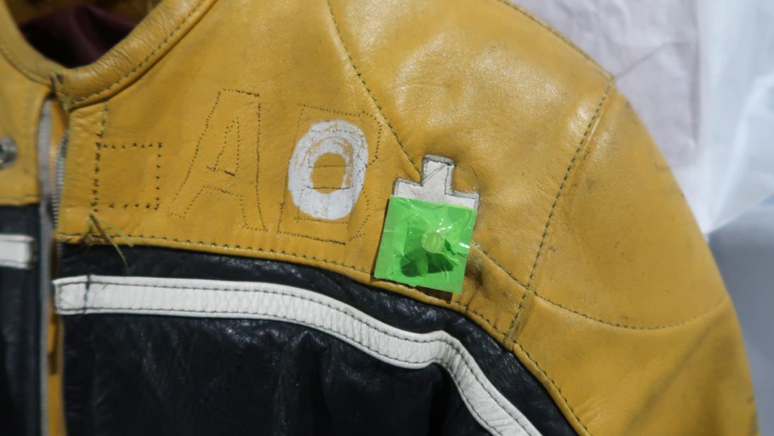 A close up of the top of a racing jumpsuit. There are white patches spelling 'O +' sewn on. There is a small magnet on top of the patch.