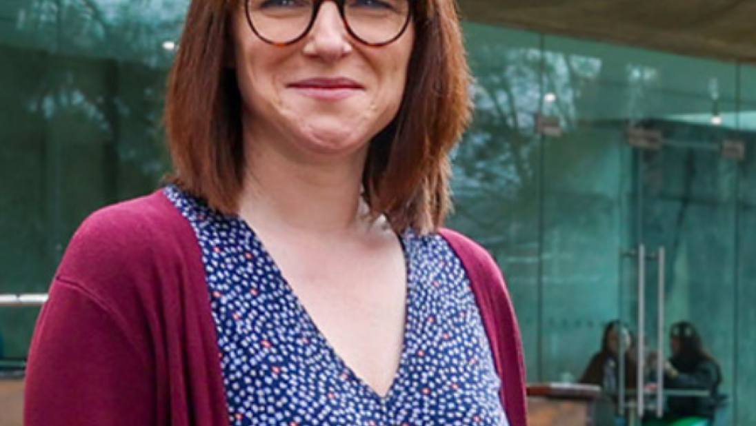 Image of a woman with short brown hair and a fringe, glasses and wearing a burgandy cardigan with a blue spotty dress