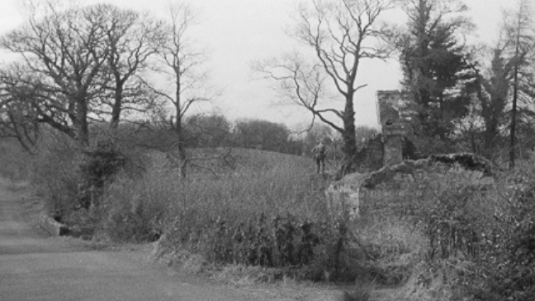 A black and white photograph of a dilapidated cottage by the side of a road.