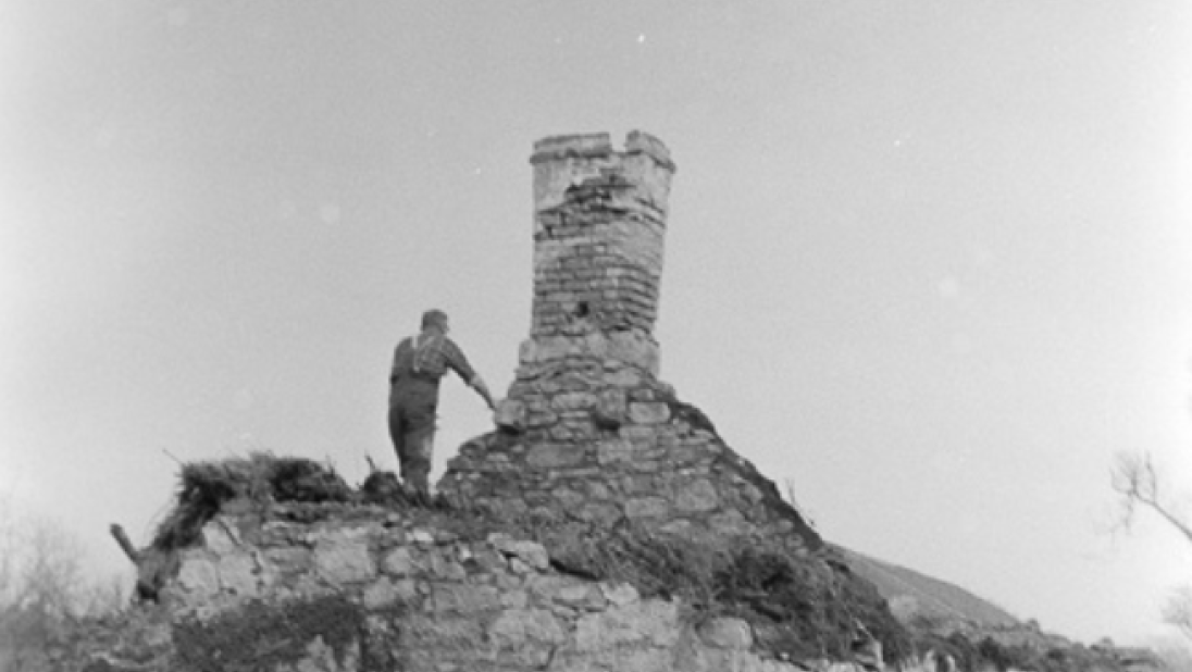 A black and white image of a man standing on a dilapidated cottage. He is holding on to the chimney, which still stands, albeit precariously. 