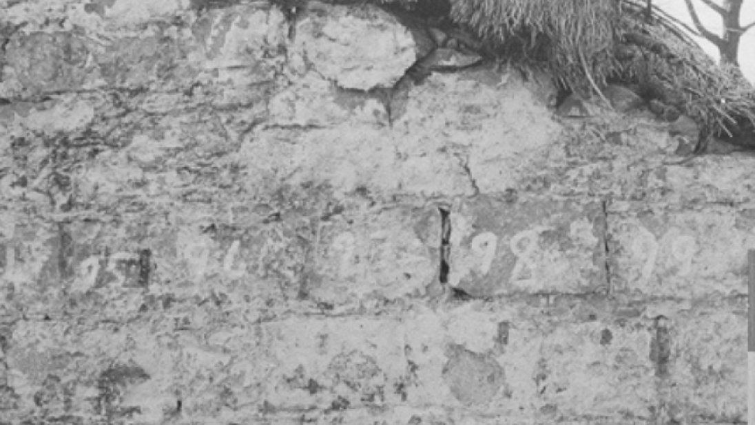 A black and white photograph of the side of a cottage wall. You can see some stones are numbered in chalk, and the slope of the thatched roof.