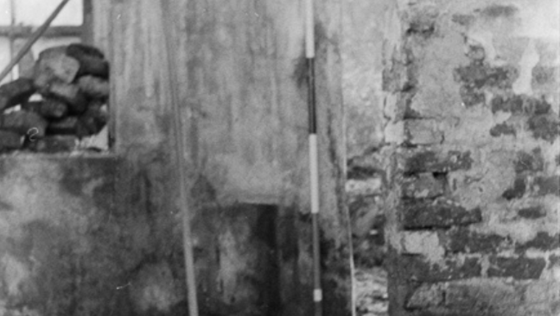 A black and white photograph of the interior of a rundown cottage. There is a large jamb wall jutting out from the right. 