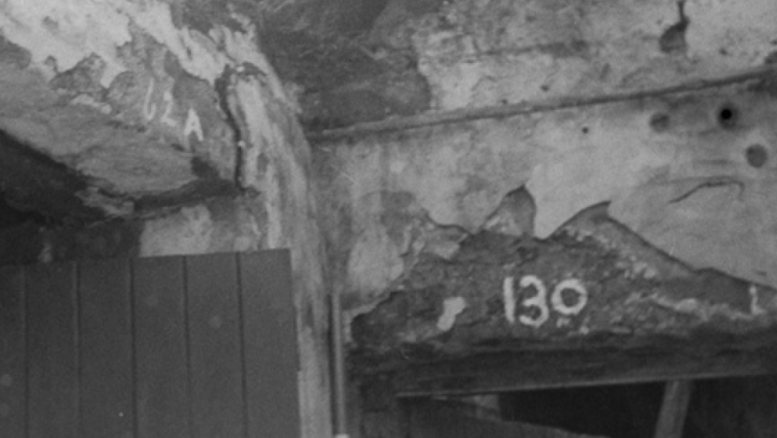 A black and white photograph of the interior of a rundown cottage. The focus is on the upper walls where two rooms ajoin. 