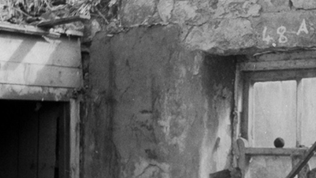 A black and white photograph of the interior or a rundown cottage showing the main room and a wall that has been raised to add more privacy. While the walls are brick, the privacy wall seems to be made of wood.