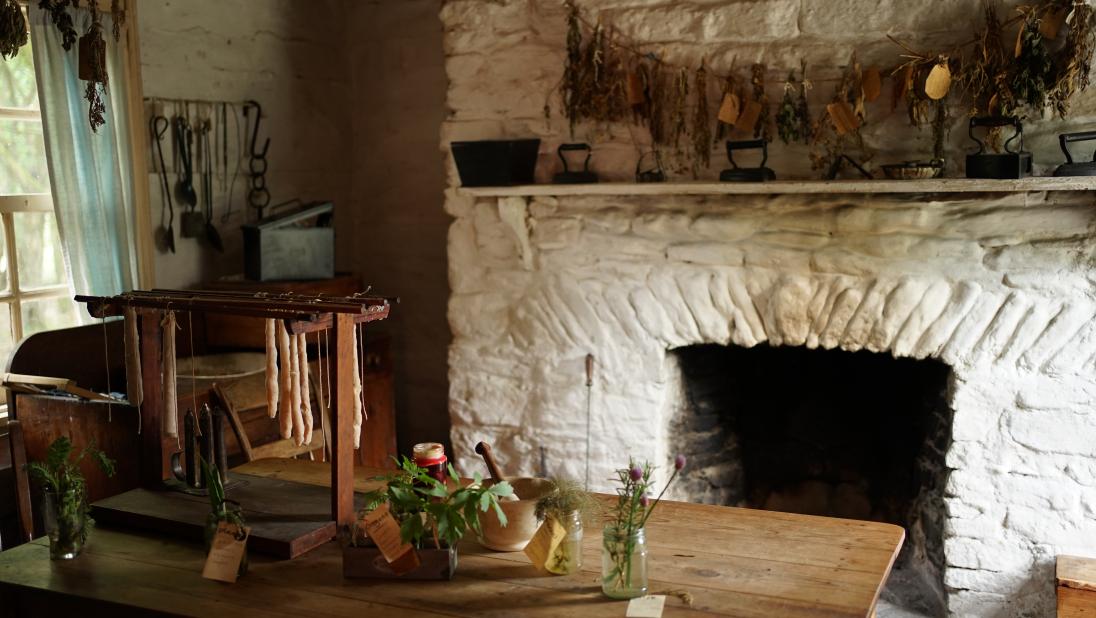 A room in a log farmhouse, with a large fireplace at the rear and a table in the centre, topped with herbs and candles setting.