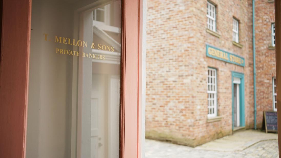 An image of the door of a bank, reading 'T. Mellon & Sons, Private Bankers'. Out the door, we can see a cobbled main street and a red brick building.