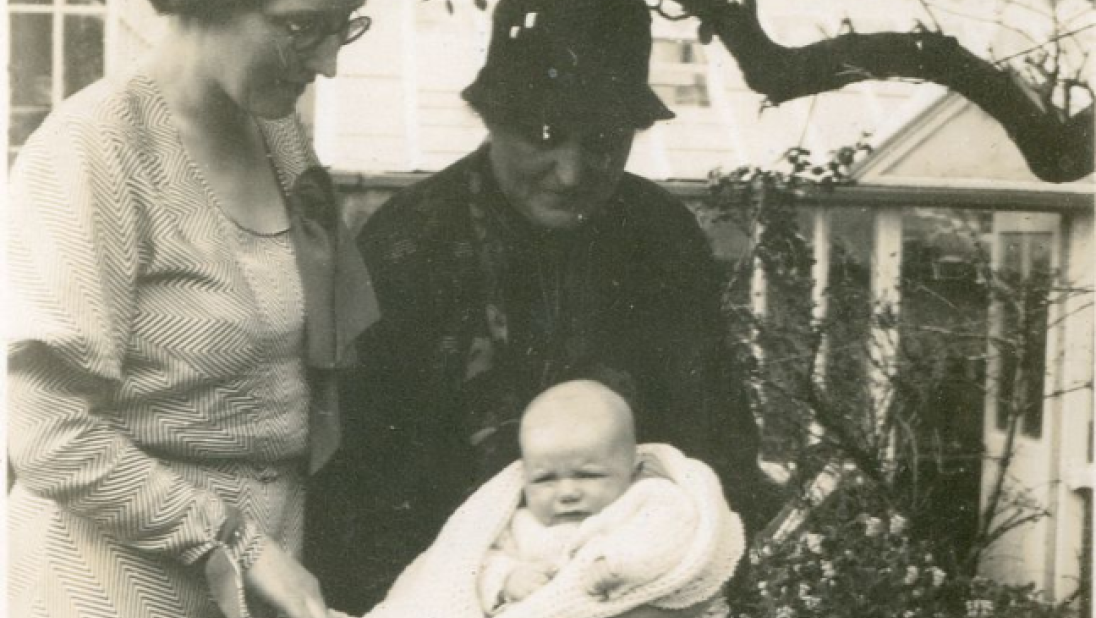 A black and white photograph of two woman, one older than the other, standing over a pram. The elder woman holds a baby in her arms, facing the camera.