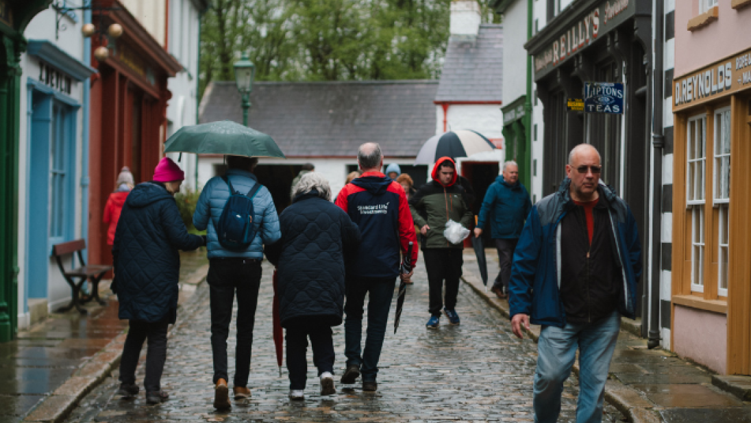 A street in Ulster American Folk Park with a crowd of people walking in the rain with an umbrella 