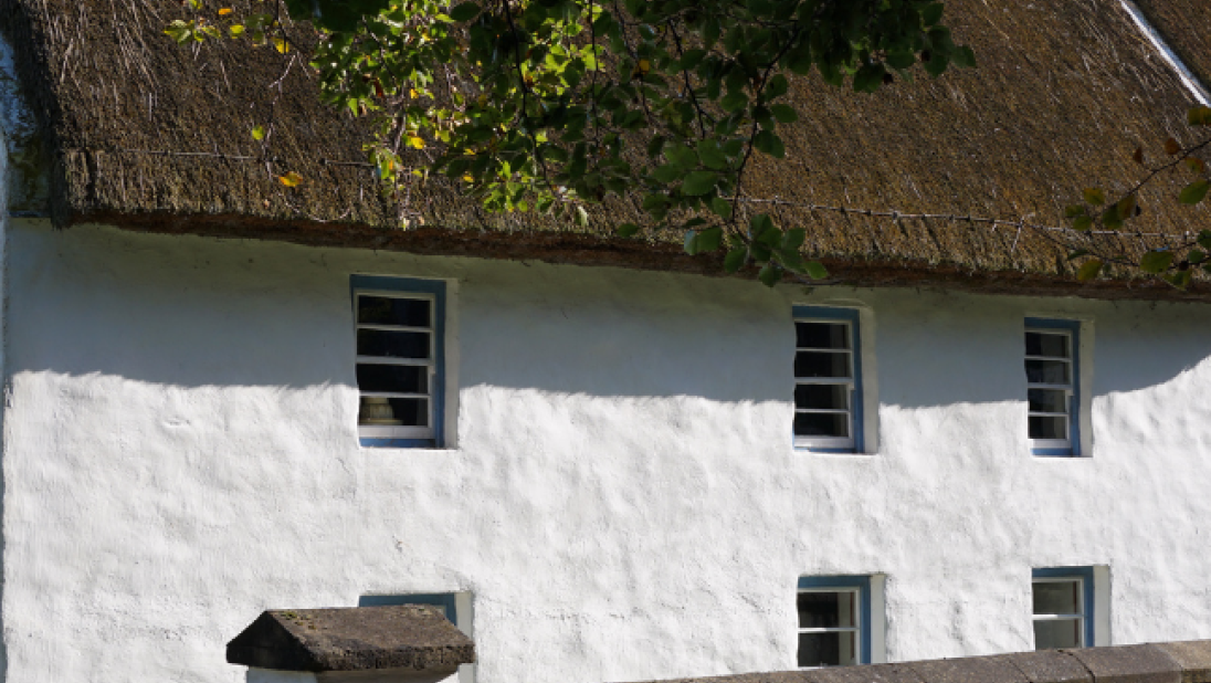 A whitewashed cottage with a thatch roof stands in the sun.