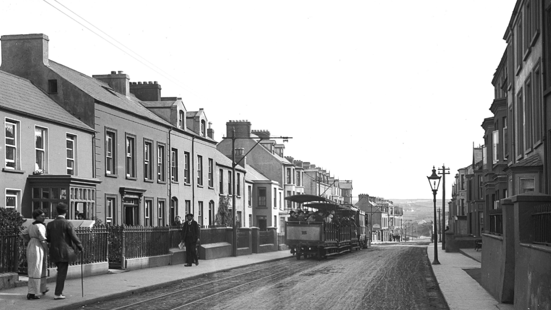 A black and white photograph showing a street with a tramline. 