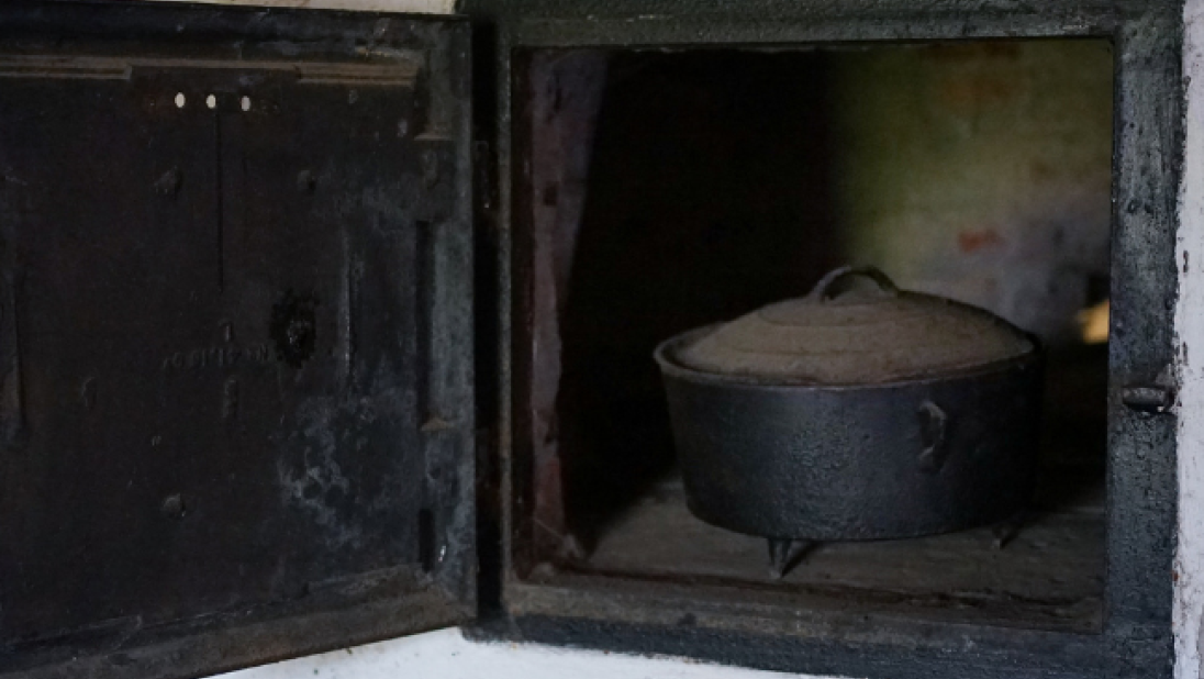 An oven set deeply into the wall is seen with its door open. An iron pot is inside.