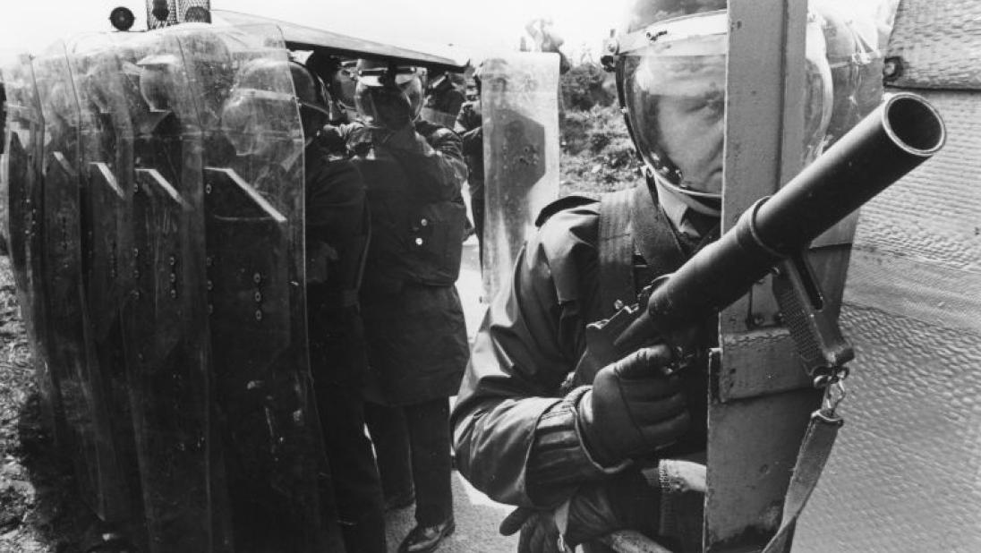 Black and white photograph of police man at rear doors of landrover with rubber bullet gun, with line of policemen behind with riot shields at funeral of hungerstriker Tom McIlwee