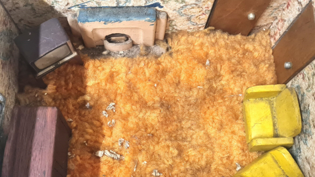 The inside of a dolls' house. The floor is a shaggy orange carpet. It is a living room, with two armchairs, a couch, and an old television in the corner.