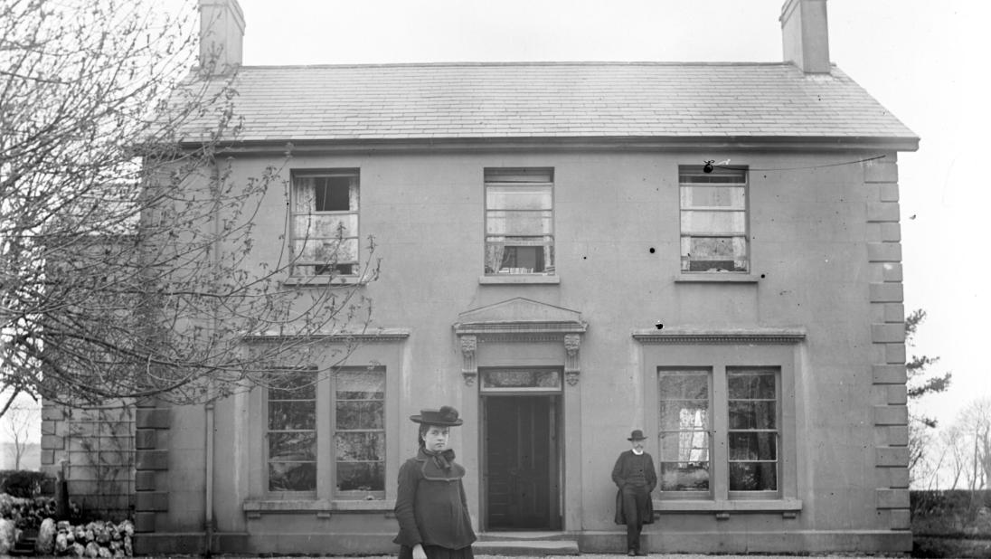 A woman stands outside a lovely two-storey home in a black and white photograph.