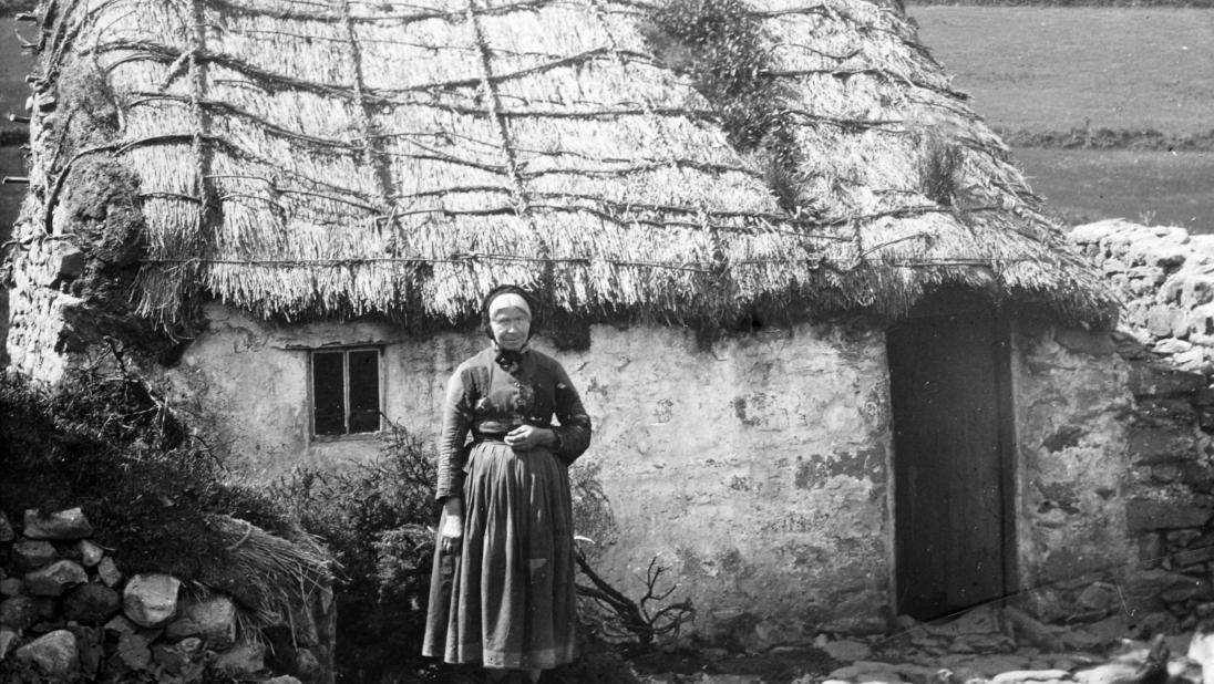 A woman stands outside a small thatched cottage in a black and white photograph.